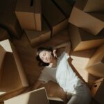Woman finds a moment of calm, meditating on the floor in a brightly lit room filled with cardboard moving boxes, symbolizi...