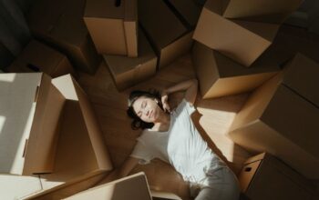 Woman finds a moment of calm, meditating on the floor in a brightly lit room filled with cardboard moving boxes, symbolizi...