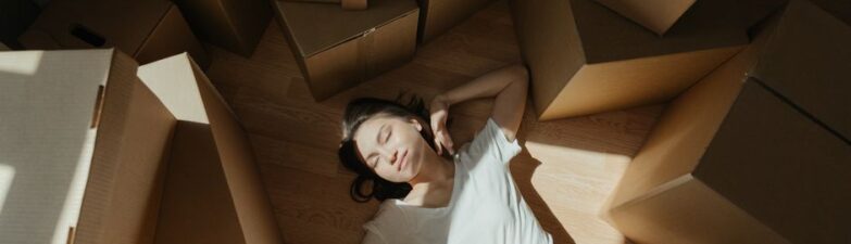 Woman finds a moment of calm, meditating on the floor in a brightly lit room filled with cardboard moving boxes, symbolizi...
