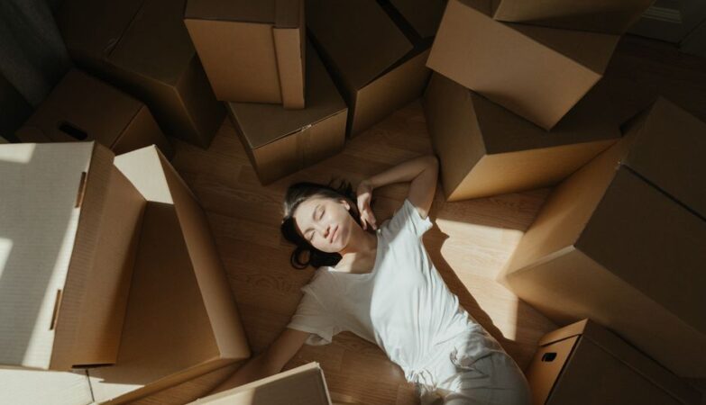 Woman finds a moment of calm, meditating on the floor in a brightly lit room filled with cardboard moving boxes, symbolizi...