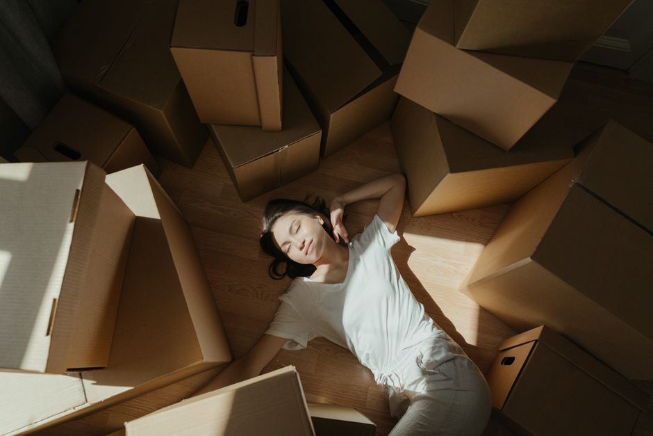 Woman finds a moment of calm, meditating on the floor in a brightly lit room filled with cardboard moving boxes, symbolizi...