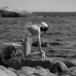 Woman in athletic wear practices yoga on a sunlit stone patio with a stunning, expansive view of the ocean and iconic rock...