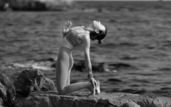Woman in athletic wear practices yoga on a sunlit stone patio with a stunning, expansive view of the ocean and iconic rock...
