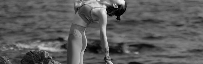 Woman in athletic wear practices yoga on a sunlit stone patio with a stunning, expansive view of the ocean and iconic rock...