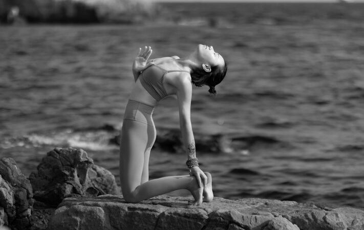Woman in athletic wear practices yoga on a sunlit stone patio with a stunning, expansive view of the ocean and iconic rock...