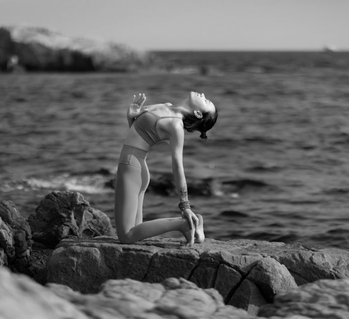 Woman in athletic wear practices yoga on a sunlit stone patio with a stunning, expansive view of the ocean and iconic rock...